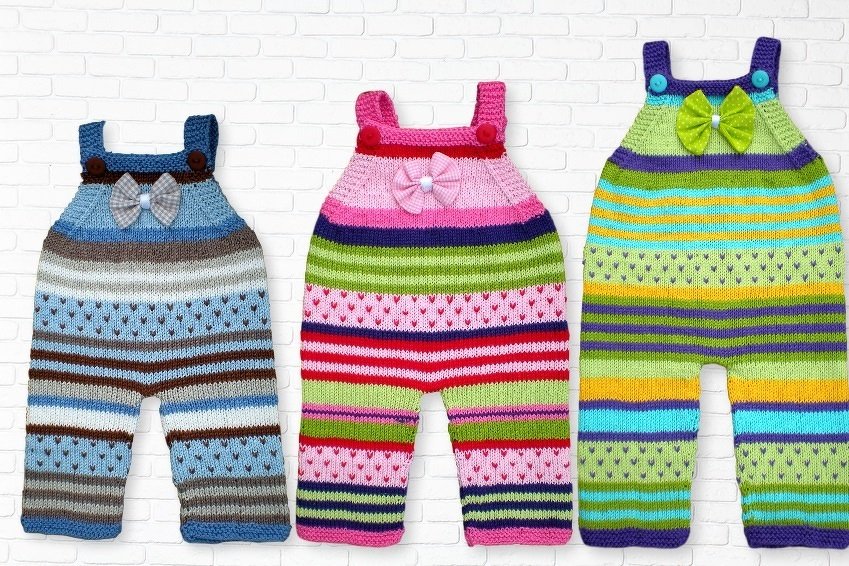 Drei handgestrickte Babystrampler mit Streifen und Herzmotiven vor weißer Ziegelwand.