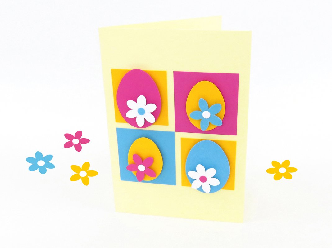 Hellgelbe Klappkarte mit vier farbigen Papier‑Eiern auf quadratischen Feldern und aufgeklebten Papierblüten, dazu verstreute Papierblumen