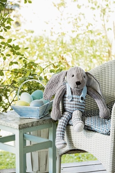 Gestrickter Amigurumi-Hase in gestreifter Latzhose auf Korbstuhl neben Korb mit pastellfarbenen Ostereiern.