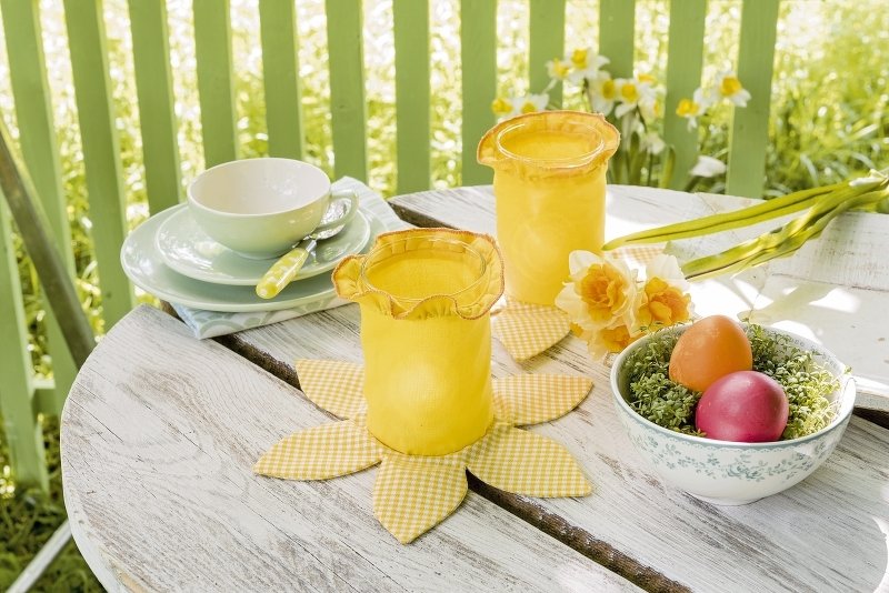 Gelbe Stoff-Windlichter auf blütenförmigen Untersetzern auf Holztisch mit Narzissen und bunten Ostereiern