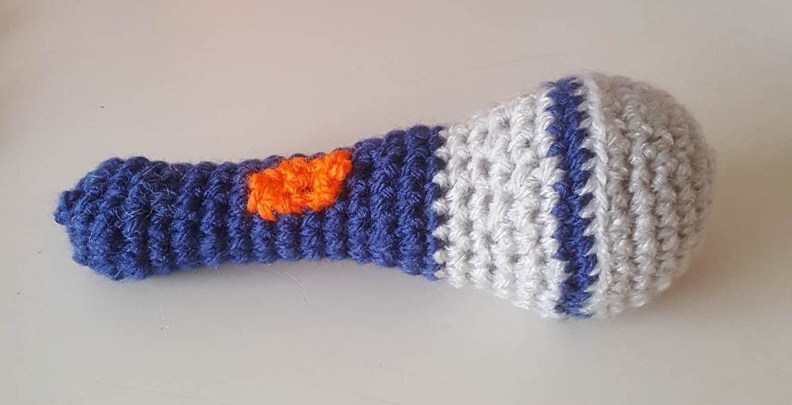 Gehäkelte Rassel liegend, blauer Griff, weißer Kopf mit blauer Linie und orangefarbener Stelle