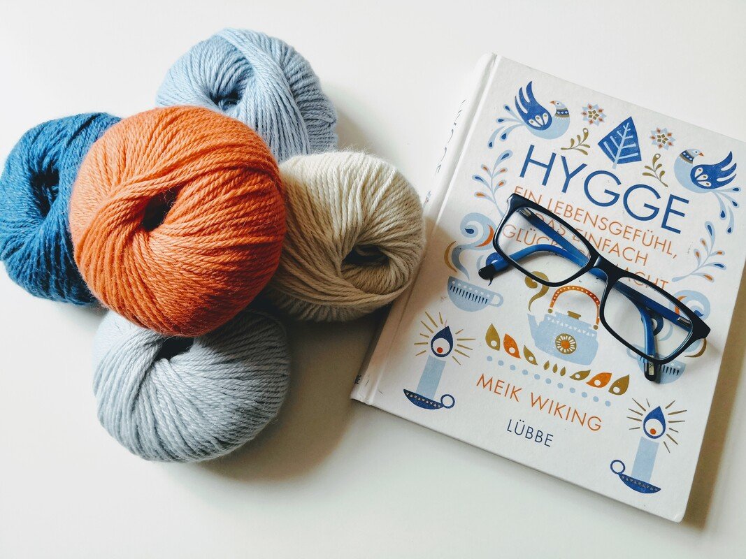 Fünf Knäuel Wolle in Blau-, Orange- und Cremetönen neben einem Buch und einer Brille.