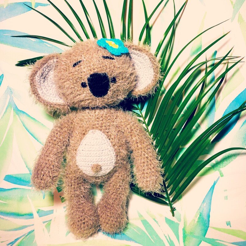Gehäkelter brauner Amigurumi-Koala mit weißem Bauchteil und blauer Blume vor Palmenblatt