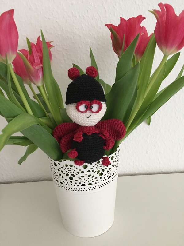 Gehäkeltes Marienkäfer-Amigurumi mit roten Flügeln im weißen Blumentopf zwischen Tulpen