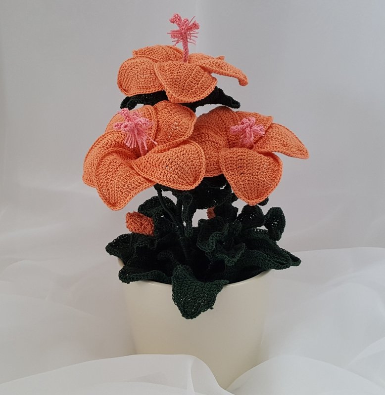 Gehäkelte Topfpflanze mit großen orangefarbenen Blüten, rosa Blütenstempeln und dunkelgrünen Blättern in hellem Topf vor hellgrauem Hintergrund.