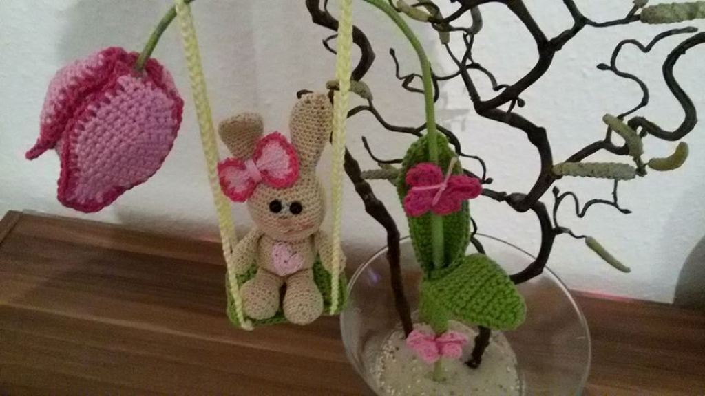 Gehäkelter beiger Amigurumi-Hase auf Schaukel mit rosa Tulpe und Schleifen an Zweig