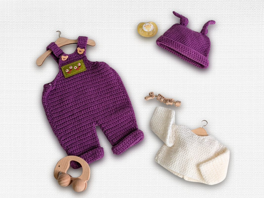Purple crocheted overalls with matching horned hat and cream cardigan for amigurumi dolls, flatlay with wooden toys