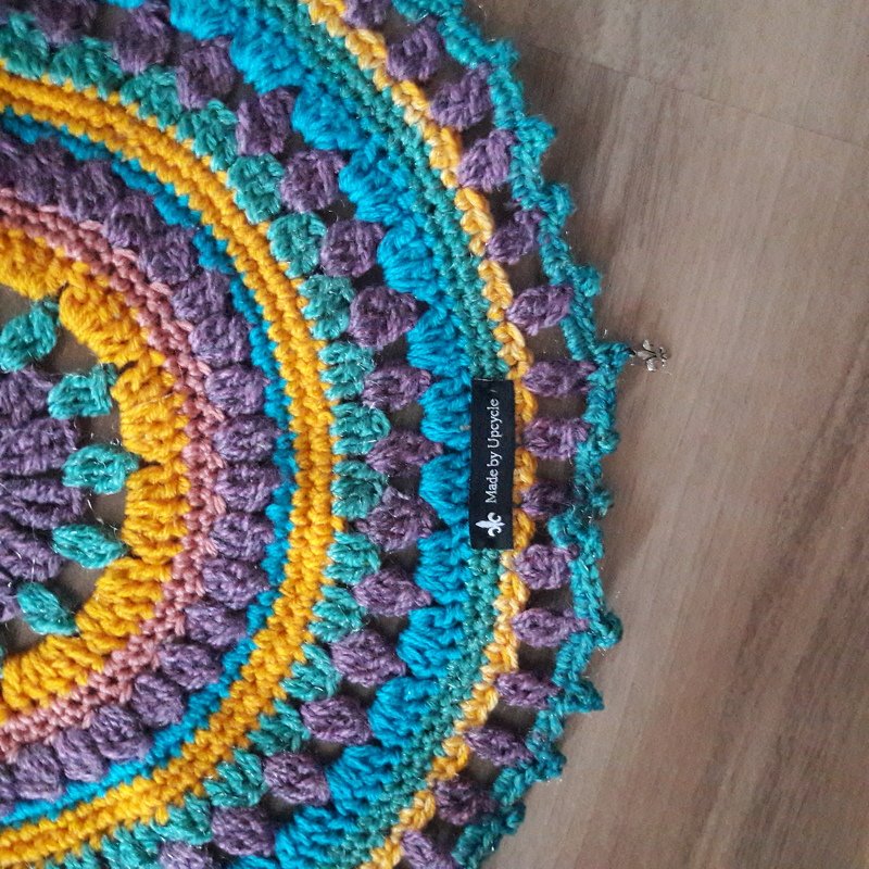 Rundes Häkel-Mandala in Türkis, Gelb und Lila mit Noppen- und Muschelmuster auf Holzuntergrund