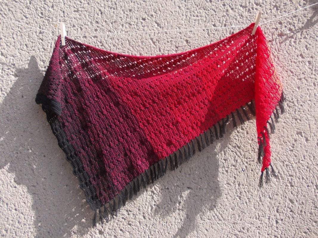 Gehäkeltes Dreieckstuch mit Farbverlauf von Schwarz zu Rot und Quasten, an einer Wäscheleine vor heller Wand hängend