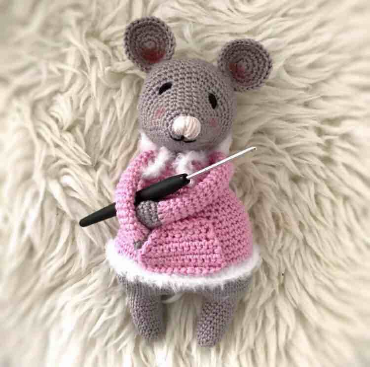 Grau gehäkelte Amigurumi‑Maus mit rosa Mantel und Häkelnadel auf flauschigem Teppich