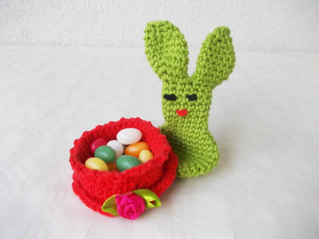 Stehender grüner Amigurumi-Hase neben kleinem roten gehäkeltem Osternest mit bunten Eiern und Stoffblüte