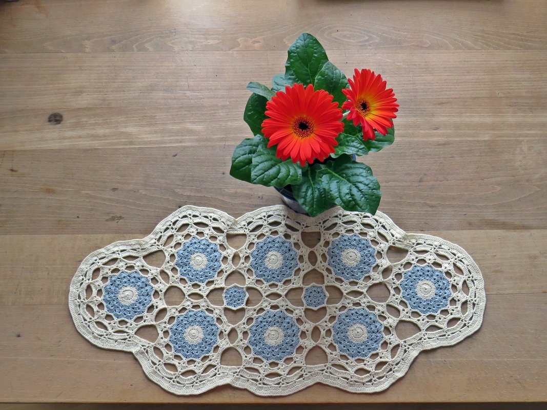 Ovales gehäkeltes Deckchen in Creme mit hellblauen Rosetten auf einem Holztisch