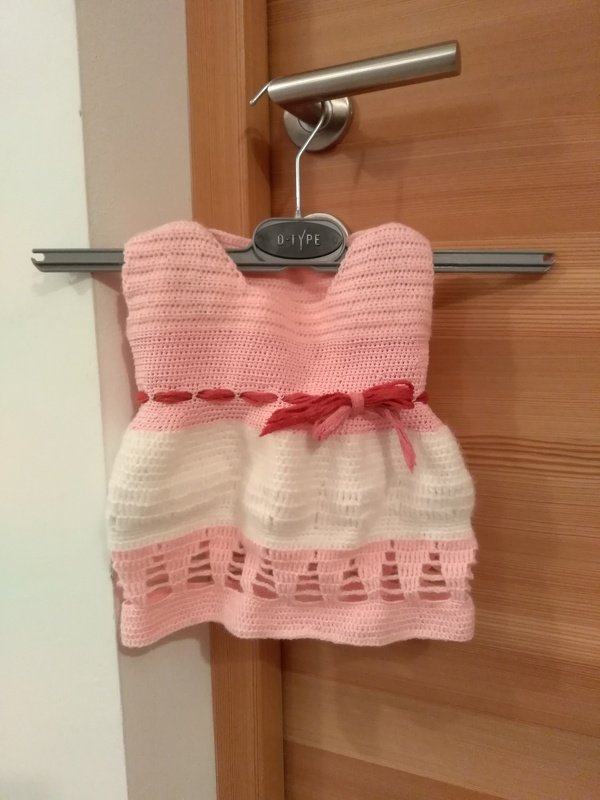 Rosa-weißes gehäkeltes Babykleid mit rosafarbenem Zierband an einem Kleiderbügel vor einer Holztür