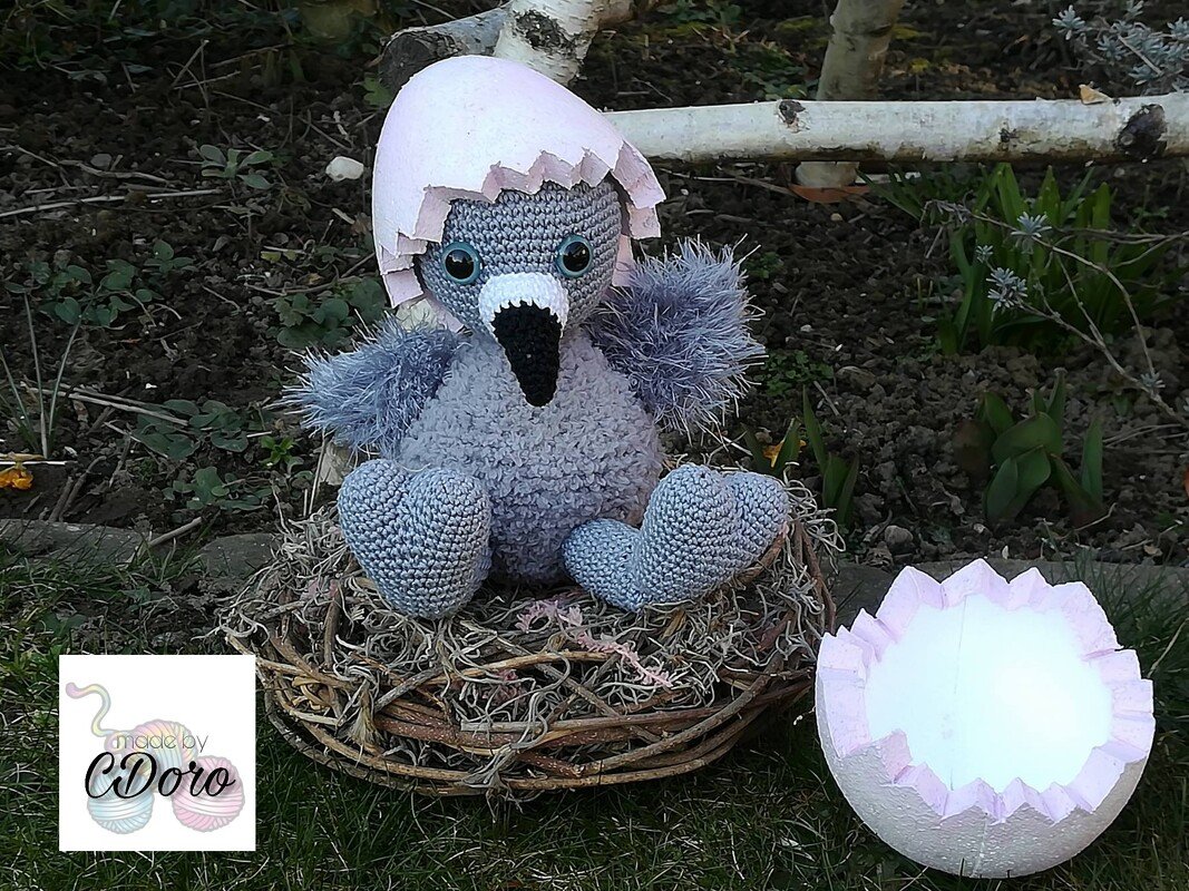 Grau gehäkeltes Küken-Amigurumi mit großen Augen und rosafarbener Eierschale im braunen Nest.