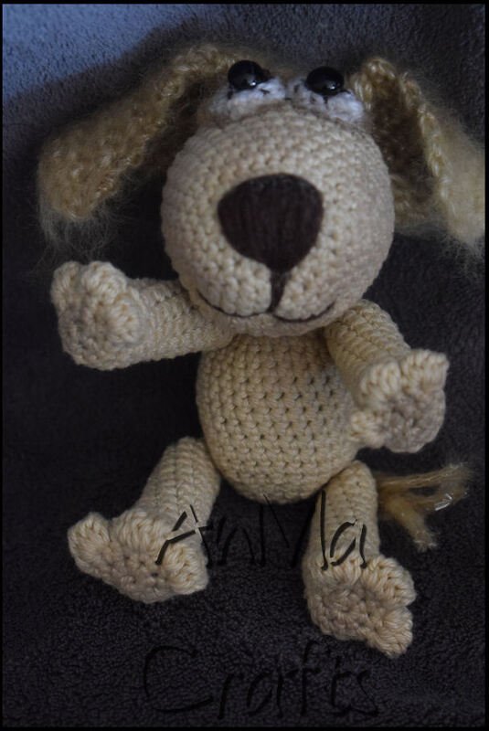 Beiger gehäkelter Amigurumi-Hund mit schlappenden Ohren und dunkler Nase auf dunklem Stoff