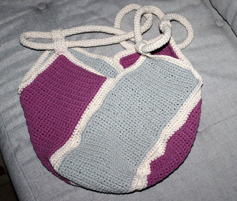 Gehäkelte Shopper-Tasche mit lila und hellblauen Farbblöcken, cremefarbener Kante und gehäkelten Tragegriffen auf grauem Sofabezug.