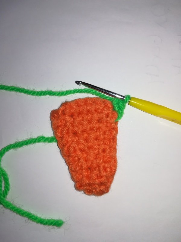 Häkelbeginn einer orangefarbenen Karotte mit grünem Blattansatz und Häkelnadel auf hellem Hintergrund