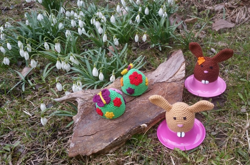 Verschiedene gehäkelte Eierwärmer mit Hasenohren und Blumenapplikationen auf Holz und Gras vor Schneeglöckchen