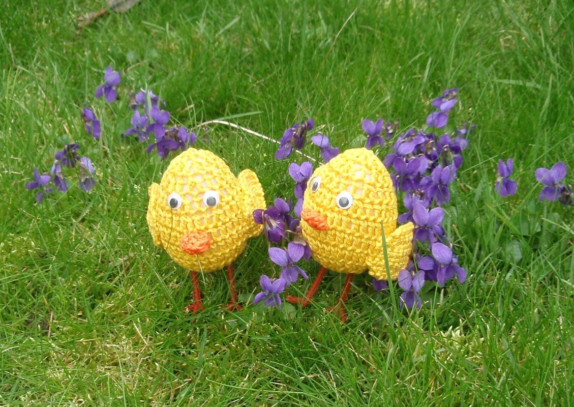 Zwei handgehäkelte gelbe Küken mit orangen Schnäbeln zwischen violetten Veilchen auf grünem Gras