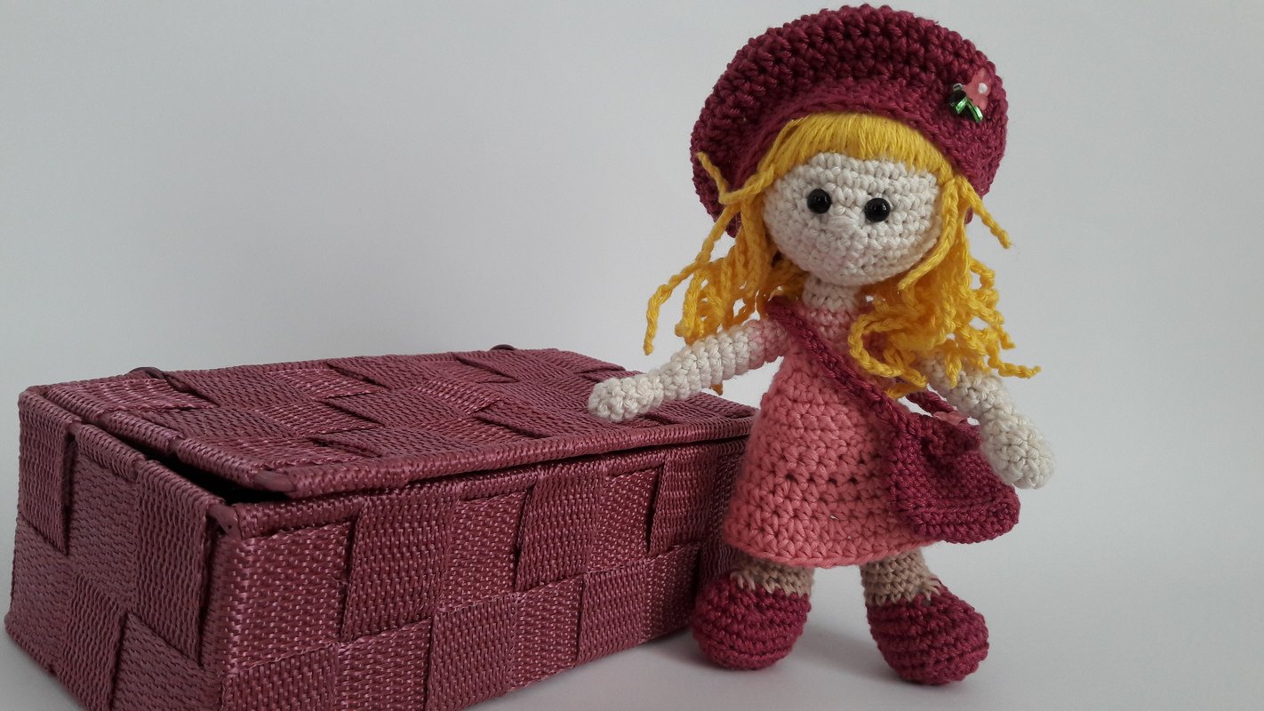Gehäkelte Amigurumi-Puppe mit blonden Wollhaaren, rosa Kleid, bordeauxfarbener Mütze, Tasche und Schuhen neben einer geflochtenen Box.