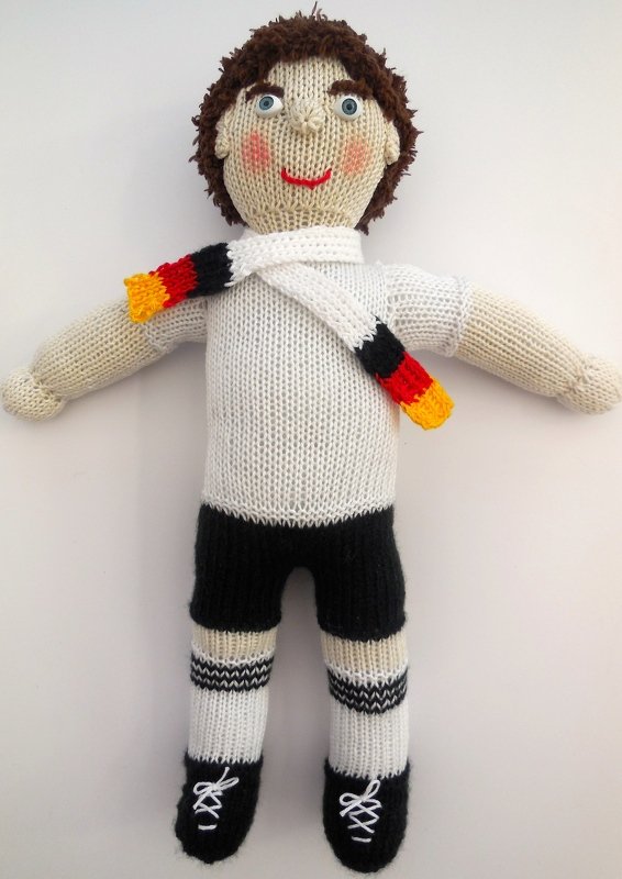 Gestrickte Amigurumi-Puppe in weißem Trikot, schwarzen Shorts, gestreiften Socken und Schal in Schwarz-Rot-Gold