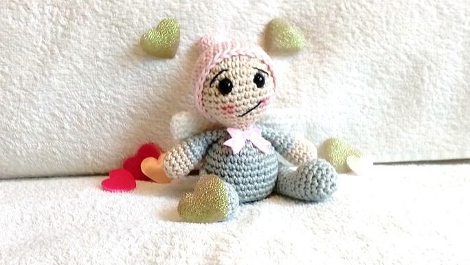 Gehäkeltes Amigurumi‑Engelchen mit rosa Mütze, weißen Flügeln und Schleifchen auf weißem Stoff zwischen Herzen.