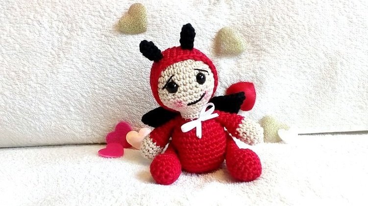 Gehäkelte Amigurumi-Glückskäfer-Puppe rot/beige mit schwarzen Fühlern und Flügeln auf weißem Stoff, umgeben von kleinen Herzen.