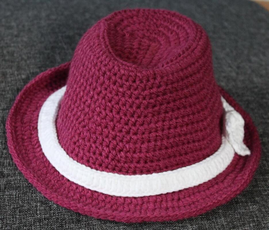 Dunkelroter gehäkelter Fedora-Hut mit weißem Band auf grauem Untergrund.
