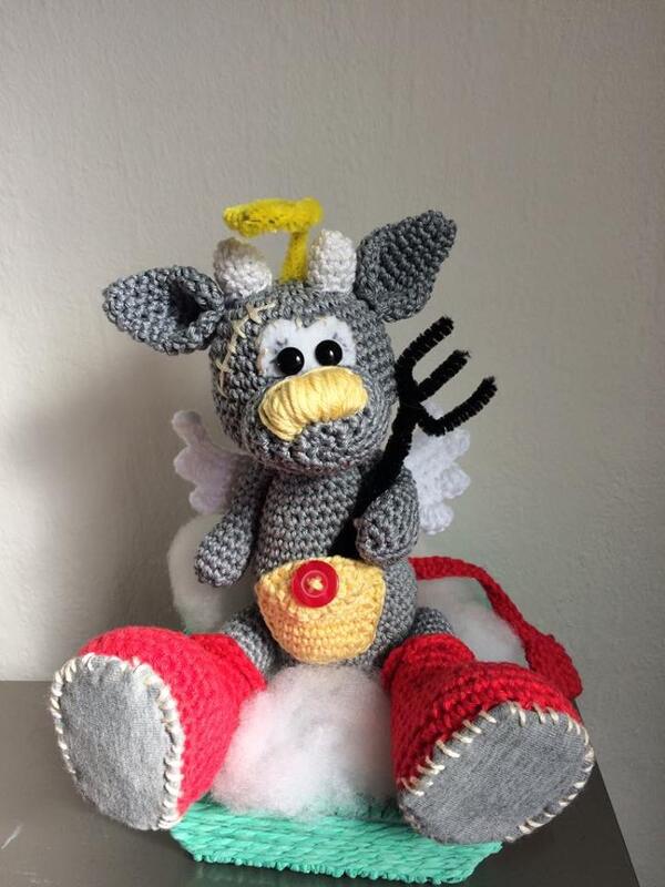 Gehäkelte graue Amigurumi-Kuh mit Heiligenschein, weißen Flügeln, gelber Schnauze, roten Stiefeln und schwarzer Dreizack-Gabel auf einer Wattewolke.