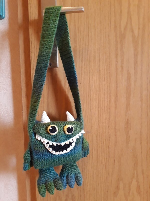 Gehäkelte Monster-Umhängetasche in Grün-Blau mit Hörnern, großen Augen und gezacktem Mund, an Türgriff hängend.