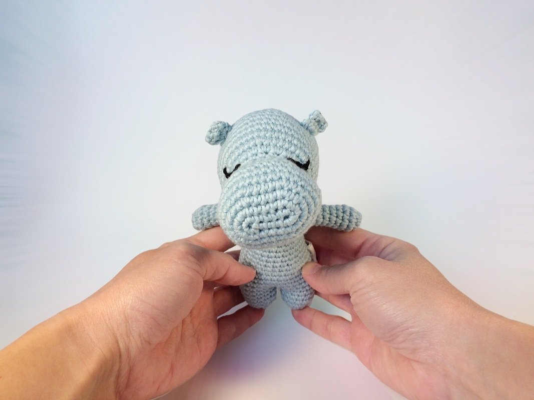 Nahaufnahme eines hellgrauen, gehäkelten Nilpferd-Amigurumis mit sichtbarer Maschenstruktur, gehalten von zwei Händen.