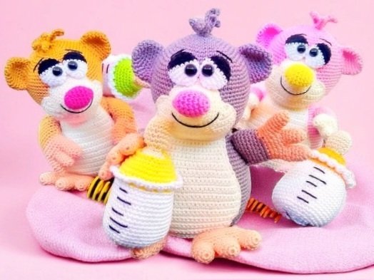 Drei gehäkelte Amigurumi-Tiere (orange, lila, rosa) mit Babyflaschen auf rosa Decke.