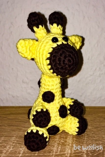 Gehäkelte sitzende Amigurumi-Giraffe in Gelb mit dunkelbraunen Flecken auf Holzuntergrund.