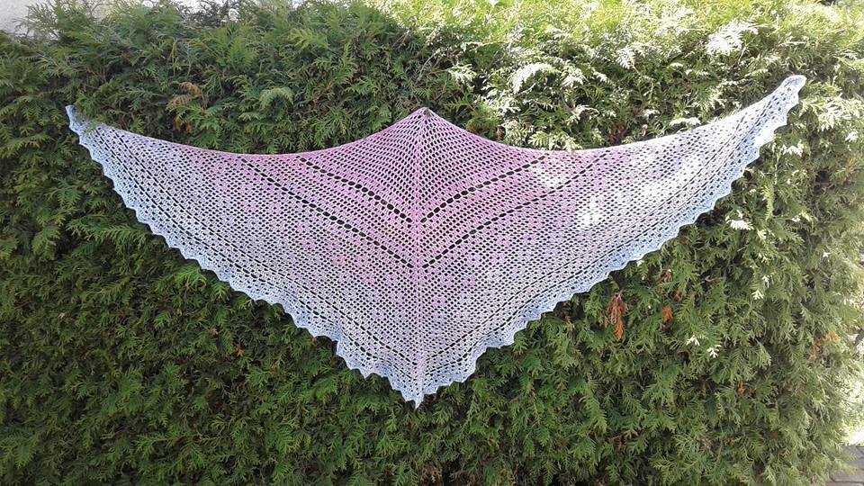 Gehäkeltes dreieckiges Spitzentuch in Pastellrosa bis Hellblau mit Muschelrand, gespannt vor einer Hecke