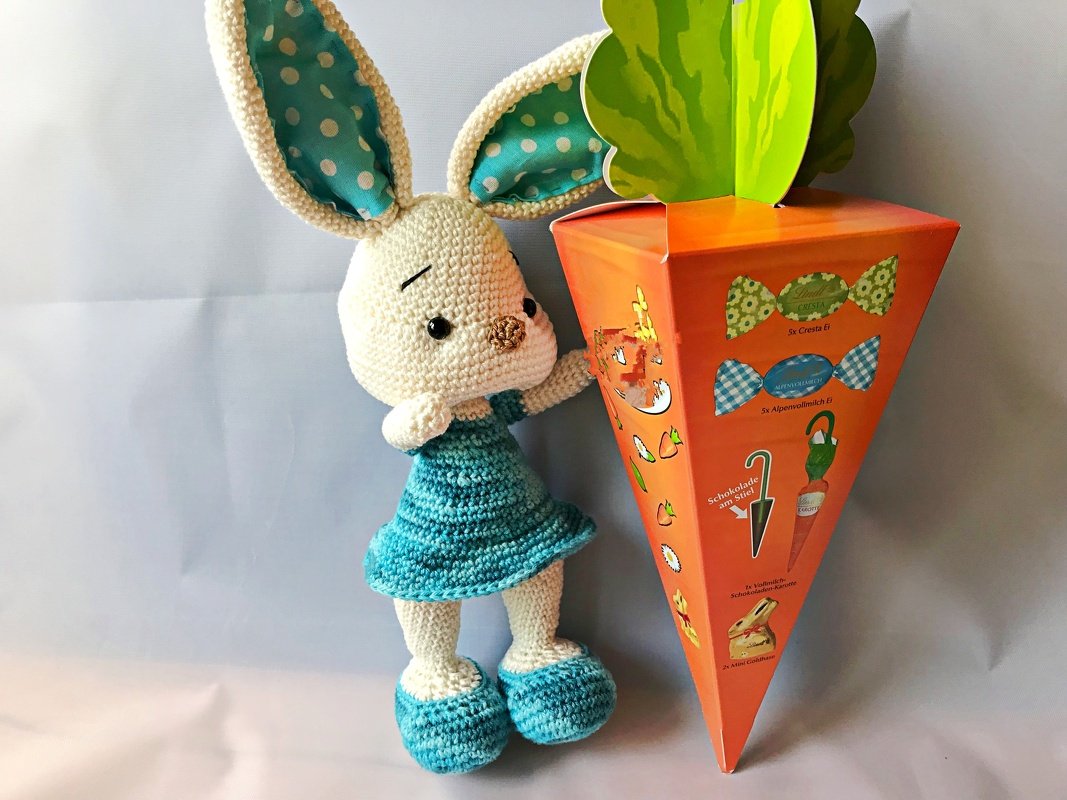 Gehäkelter Amigurumi-Hase mit türkisfarbenem Kleid neben orangefarbener Karottenbox