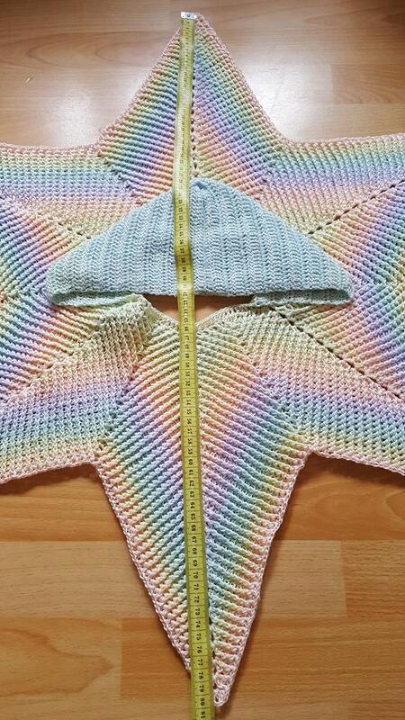 Gehäkelte sternförmige Baby-Wickeljacke in Pastellregenbogen mit hellblauer Mütze und Maßband auf Holzfußboden