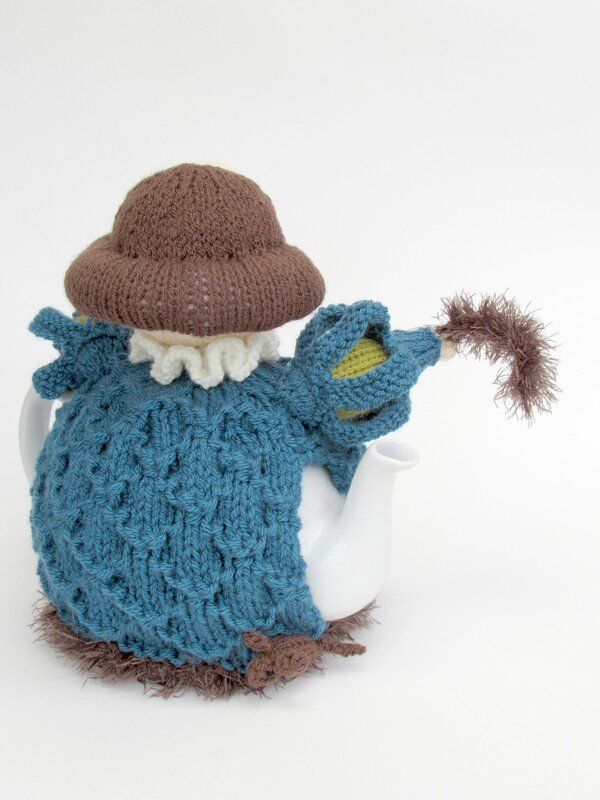 White teapot dressed in a whimsical blue knitted cozy with a brown hat and ruffled collar