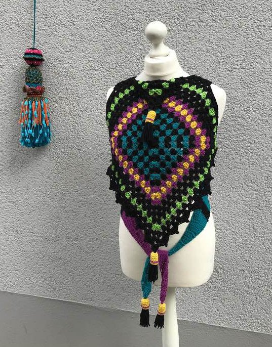 Gehäkeltes schwarzes Poncho-Top mit bunten Granny Squares und Quasten auf einer Schneiderpuppe vor grauer Wand.