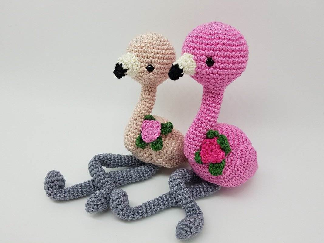 Zwei gehäkelte Amigurumi-Flamingos in Rosa und Beige mit grauen Beinen und Rosenapplikationen