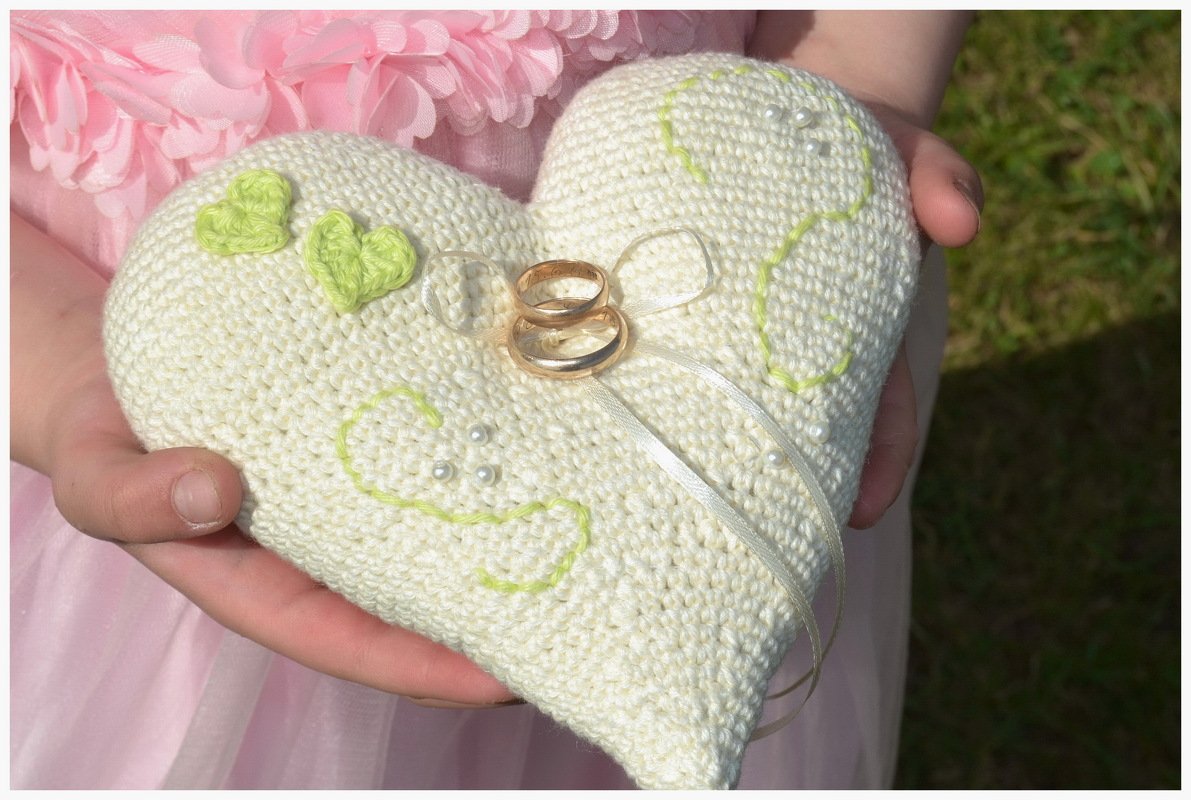 Gehäkeltes cremefarbenes Herz-Ringkissen mit Trauringen, grünen Häkelherzen und Perlen, gehalten von Händen.
