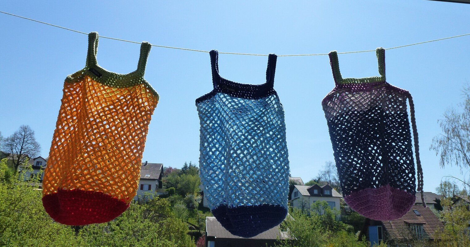 Drei gehäkelte Netzbeutel in bunten Farbkombinationen an einer Wäscheleine vor blauem Himmel.