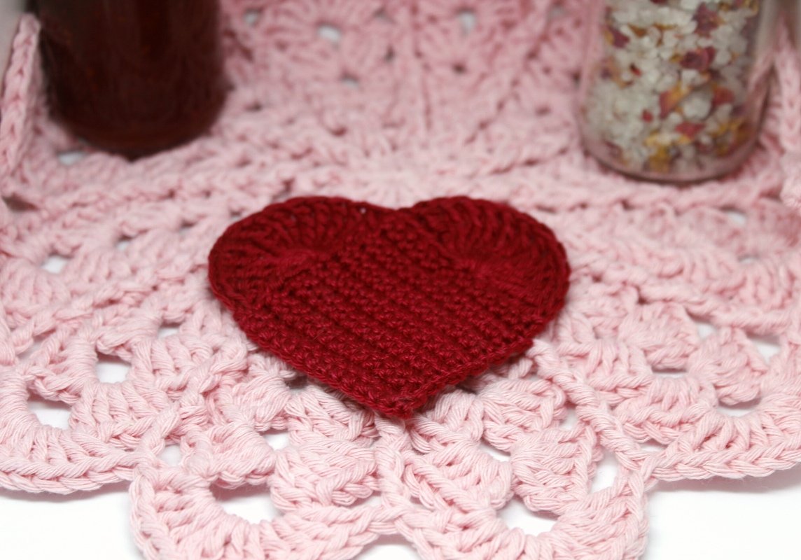 Handgehäkeltes rotes Herz auf einem hellrosa Häkeldeckchen mit detailreicher Musterung.