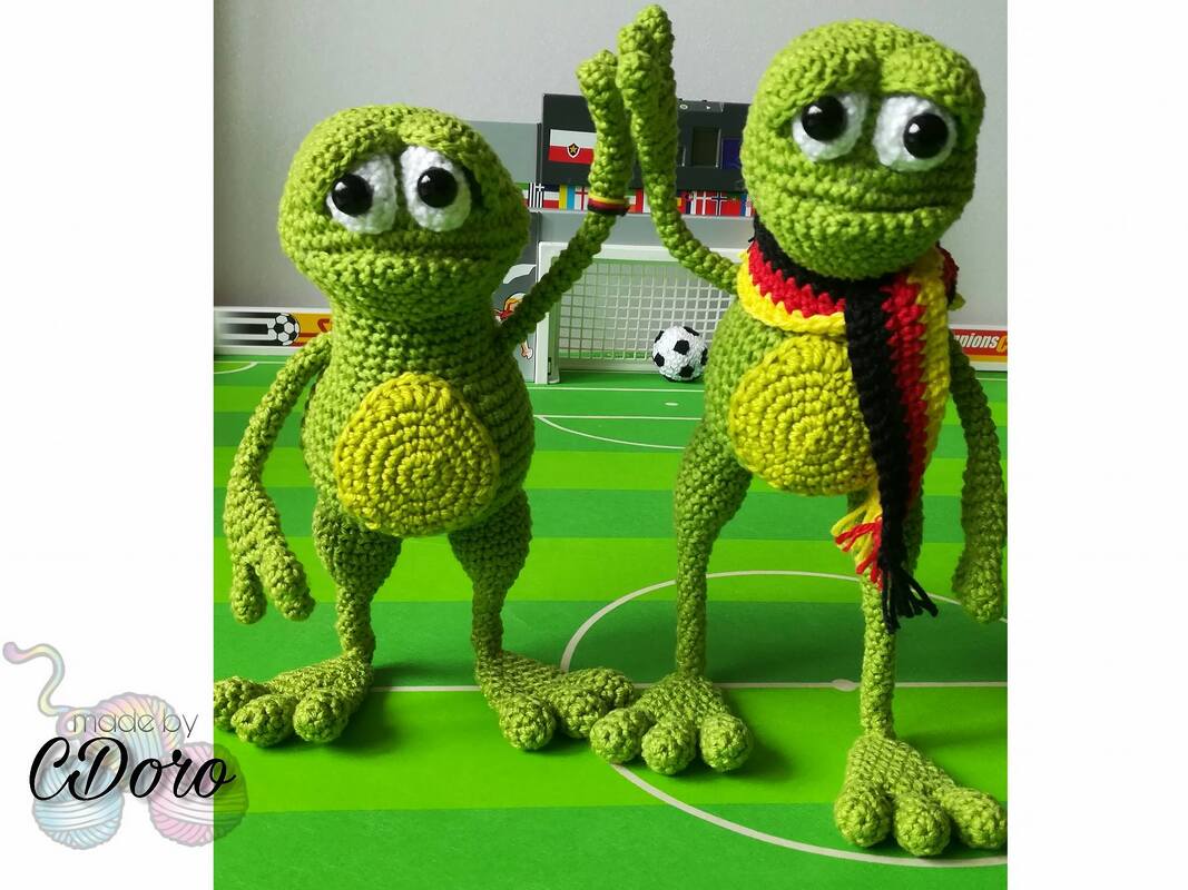 Two green crocheted frog amigurumi on a miniature soccer pitch, one wearing a striped scarf.
