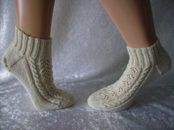 Cremefarbene, gestrickte Knöchelsocken mit Zopf- und Ajourmuster an Modellfüßen.