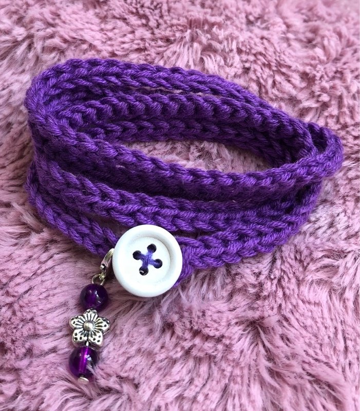 Gehäkeltes violettes Wickelarmband mit weißem Knopf und Perlen-Blumen-Anhänger auf rosa Plüsch