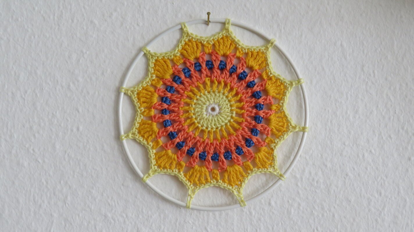 Häkel‑Mandala im weißen Ring mit gelben, korallenfarbenen und blauen Garnrunden an heller Wand