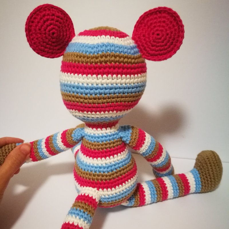 Rückansicht eines gestreiften, gehäkelten Amigurumi-Affen; eine Hand hält einen Arm, große runde Ohren sichtbar