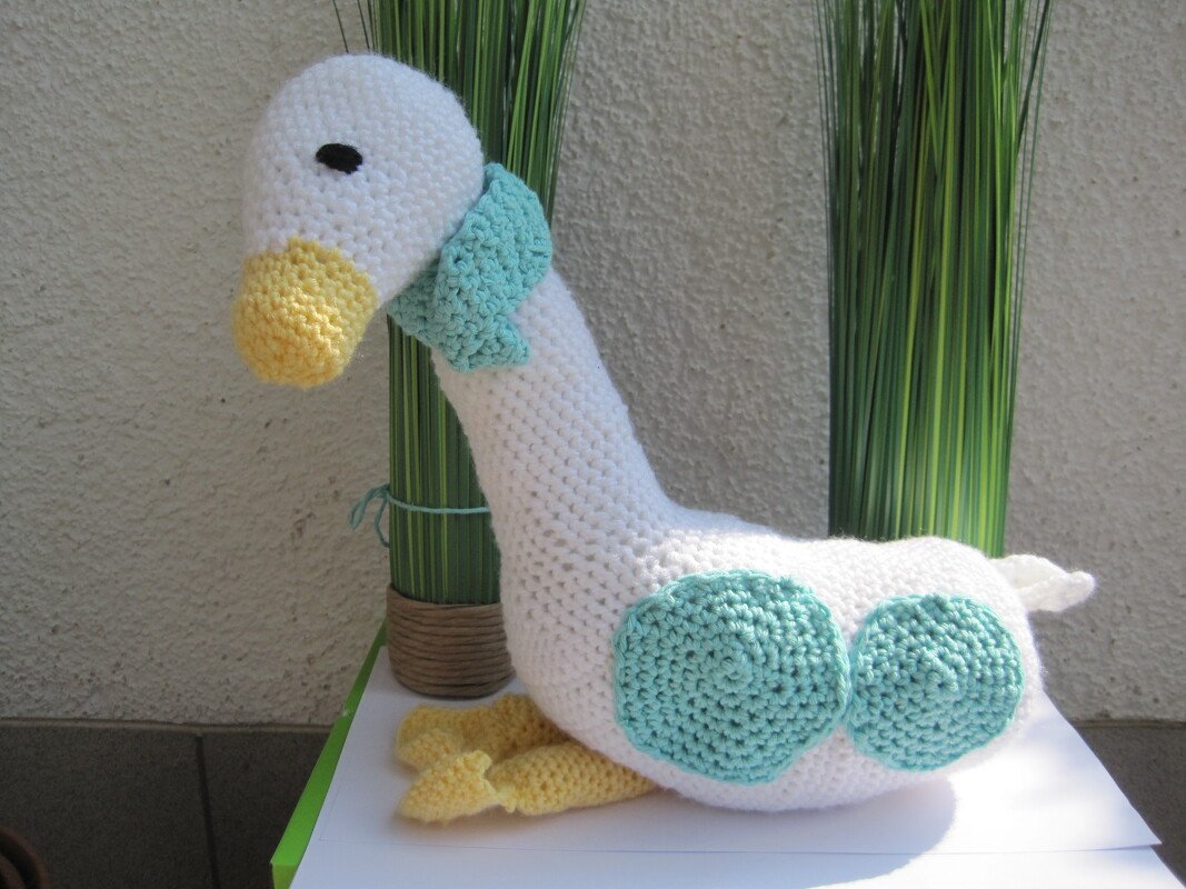 Gehäkelte weiße Amigurumi‑Gans mit gelbem Schnabel und Füßen, türkisfarbenen Kreisen und Halstuch