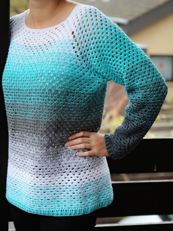 Woman wearing a long-sleeve crocheted sweater with ombre colors in white, teal, and gray.