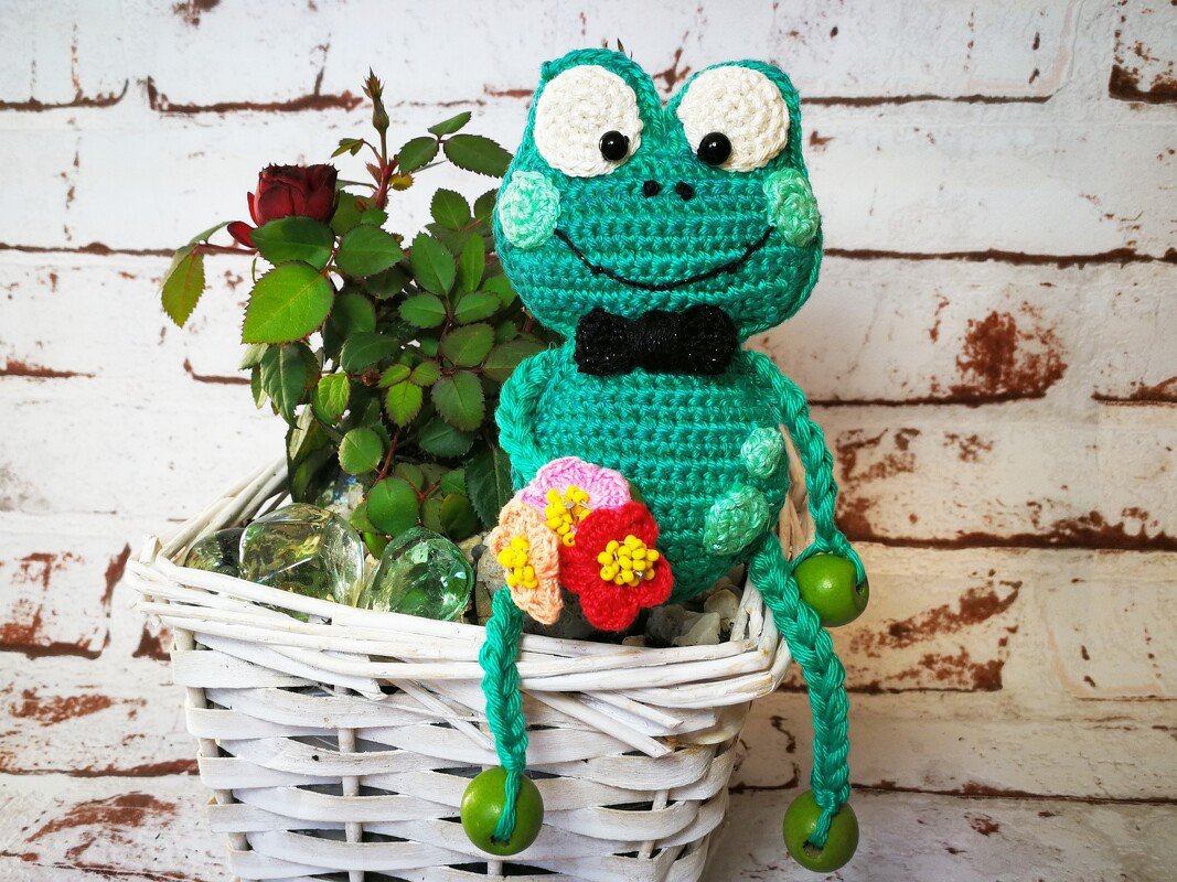 Grüner, gehäkelter Amigurumi-Frosch mit schwarzer Fliege und Häkelblüten auf weißem Korb neben einer roten Rose.